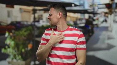 Çizgili gömlekli genç adam şehir terasında boynuna dokunuyor açık havada oturma ve hareketli şehir arkaplanı gün boyunca rahatsız görünüyor..