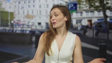 Beyaz kolsuz bluzlu kadın, yoğun şehir caddesinde gün ışığında elleri yanında seni çağırıyor; hoş geldin oyunculuğu daveti.