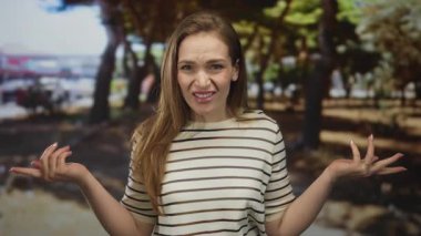 Çizgili gömlekli kadın, parlak güneş ışığı altında meşe ağaçlarının yanındaki yeşil parkta el ve kırışıklıklar silkeliyor..