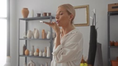 Kadın, kilden vazolar ve raflarla çevrili bir çömlek atölyesinde dudaklarına akıllı telefon tutuyor..