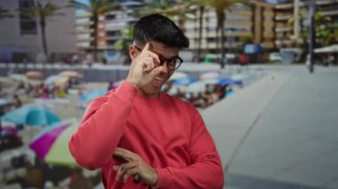 Sahilde gülümseyen gözlüklü genç adam canlı bir sahil arka planı ve palmiye ağaçları, kırmızı bir süveter giyiyor, açık havada neşeli bir hava var..