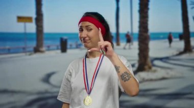 Madalyalı bir kadın güneşli sahil yürüyüşünde saç bandı ve dövme takıyor, başarıyı ve kendine güveni somutlaştırıyor..