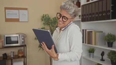 Kısa saçlı ve gözlüklü bir kadın elinde tablet, omuzda akıllı telefonu raflar ve bitkilerle dengeliyor. Mutluluk..