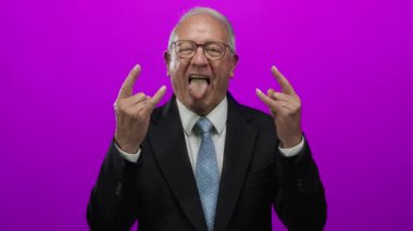 Takım elbiseli son sınıf öğrencisi, parlak pembe arka plan duvarına karşı neşeli bir ifadeyle taş el hareketleri yapıyor..