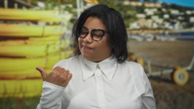 Beyaz tişörtlü ve gözlüklü, iri yarı, Latin bir kadın sahil kenarındaki arka plana doğru başparmağıyla güvenle duruyor..