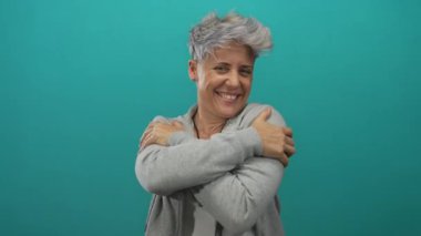 Gülümseyen kısa saçlı kadın elleriyle omuzlarını sarmalıyor. Stüdyo duvarına yaslanıyor..