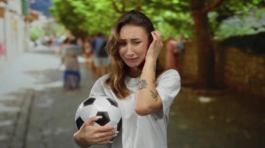 Futbol topu tutan genç bir kadın gündüzleri şehir sokaklarında duygusal görünür. Sıradan stil ve dövme sanatı sergiler..