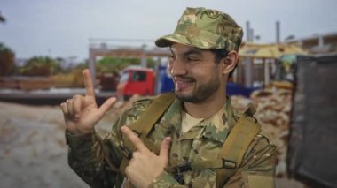 Kamuflaj üniformalı bir adam inşaat alanında parmağını öne doğru gösteriyor. Sırt çantası takıyor. Gurur görevi..