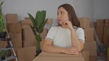 Genç bir kadın evdeki kutular arasında elleri çenesinde karton kutunun üzerinde oturur; sessiz tefekkür.