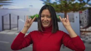 Kırmızı kazaklı bir kadın, taştan bir jest yapıyor. İki elini de şehir sokaklarında gezdiriyor. Canlı ve genç bir hava katıyor..