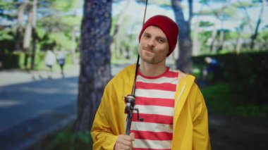 Sarı ceketli ve elinde olta tutan kırmızı bereli genç adam arka planda bulanık ağaçlarla parkta gülümsüyor, boş zamanlarını yakalıyor..