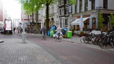 Bisikletli bulanık Amsterdam Caddesi, yürüyen insanlar ve açık hava kafeleri yumuşak akşam ışığı altında canlı bir şehir manzarası yaratıyor..