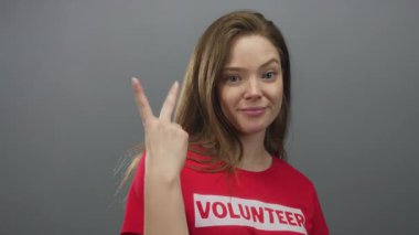 Kırmızı gönüllü gömleği giyen kadın gri stüdyoda zafer işareti yapıyor; gönüllülük cemiyeti kendine güveni destekliyor.