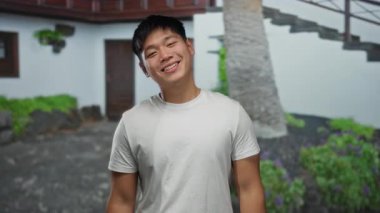 Neşeli bir ruh hali içindeki genç adam canlı bir şehir caddesinde gözlerini kapar ve yeşil bir şehir arka planında neşeli bir yüz ifadesi sergiler..