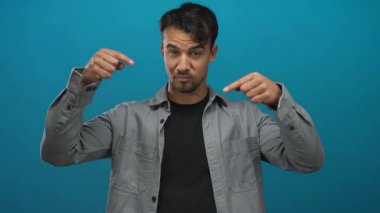 Latin kökenli genç bir adam mavi bir arkaplana işaret ediyor, yönünü vurguluyor ve gündelik bir ortama odaklanıyor, yönlendirme ve yönlendirme amacıyla ideal..