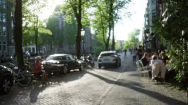 Avrupai bir şehirde insanların dışarıda yemek yediği, patikada ağaçların sıraya girdiği ve kaldırım kenarına park edilmiş arabaların olduğu bulanık bir sokak manzarası..