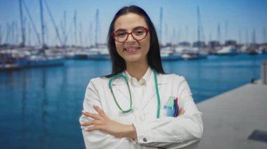 Steteskoplu İspanyol kadın doktor güneşli bir günde arka planda mavi su ve botlarla marinada güvenle gülümsüyor..