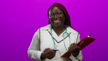 Gözlüklü ve steteskop takan Afrikalı Amerikalı kadın gülümsüyor, izole edilmiş pembe arka plan üzerine bir pano uzatıyordu..