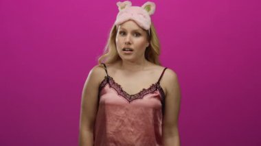 Pembe gecelikli genç sarışın kadın, canlı pembe bir arka plana karşı uyku maskesiyle gülümsüyor, neşeli ve rahat bir ruh hali yakalıyor..