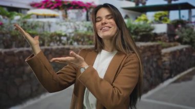 Genç İspanyol kadın gülümsüyor ve elleri ile canlı bir şehir caddesinde etrafı renkli çiçekler ve açık hava dekorlarıyla çevrili öğleden sonra ışığında şık bir ceket giyiyor..
