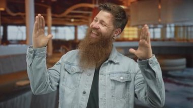 Çalı sakallı, kot ceket giyen bir adam iki elini de binanın içinde açıyor; coşku.