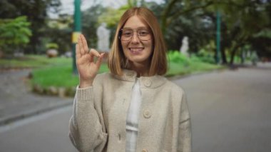 Omuz hizasında saçları olan genç sarışın kadın park yerinde sağ elle işaret dilini gösteriyor; onay pozitiflik anlaşması iyimserliği.