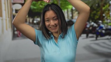 Mavi tişörtlü kadın şehir sokaklarında elleri ile kalp hareketi yapıyor. Arka planda bulanık motosikletler ve ağaçlar olan şehir arka planında gülerek gülümsüyor..