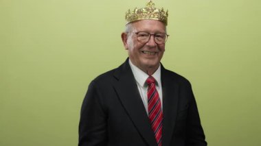 Takım elbiseli ve taç giymiş kıdemli bir adam, başparmağıyla güvenle gülümsüyor, parlak sarı bir arka plana işaret ediyor, mizahi ve asil bir iş konseptini somutlaştırıyor..