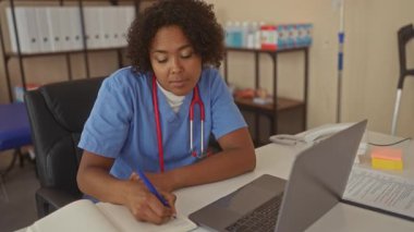 Not defterine mavi kalemle açık bir dizüstü bilgisayarın yanına notlar yazan bir kadın..