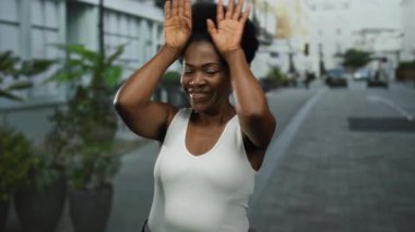 Gülümseyen bir kadın, şehir sokaklarında elleri ile neşeli bir jest yaparak, açık şehir ortamında neşe ve pozitifliği canlı bir enerjiyle vurguluyor.