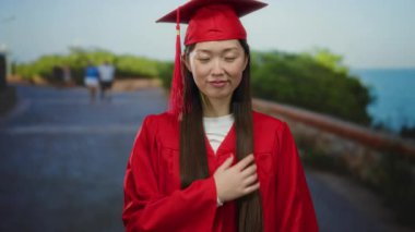 Kırmızı mezuniyet elbisesi giyen kadın deniz kenarında uzun saçlı ve arka planda yeşillikli..