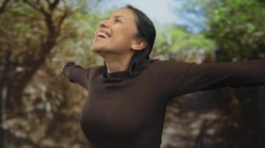 Yeşil bir park ortamında neşeli bir şekilde gülümseyen kadın, sakin bir ifadeyle doğayı kucaklayan, güneşli gökyüzünün altında mutluluk ve huzur gösteren bir kadın..