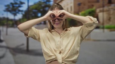 Çıplak kollarıyla dürbün oluşturan bir kadın, güneşli bir şehir sokağında binalarla dolu bir jest yapıyor, neşeyi keşfediyor..