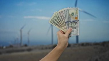Arka planda yel değirmenleri ve yeşil yatırımları ve çevresel karları temsil eden yenilenebilir enerji ve ekonomik başarıyı sembolize eden yel ele tutuşan Amerikan parası