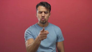 Mavi gömlekli genç İspanyol adam, izole edilmiş kırmızı arka plana ciddi şekilde parmak işareti yapıyor. Kararlılık ve odaklanma gösteriyor.