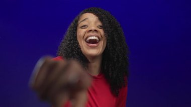 Mavi stüdyoda, kıvırcık saçlı, kırmızı bluzlu ve gülen yüzlü bir kadın kamerayı işaret ediyor..
