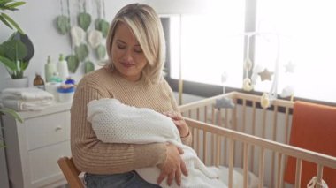 Sarışın kadın, güneşli bir yatak odasında elleri beyaz bir battaniyeye sarılı yeni doğmuş bebeği nazikçe kucaklar..