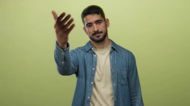 Genç adam, parlak sarı arka plana karşı davetkar bir şekilde elini uzatır. Günlük kıyafetler giyer ve arkadaşlığını ifade eder. Parlak, izole bir ortamda hoş geldin jesti yapar..