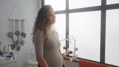 Hamile kadın parlak bir yatak odasında duruyor. Karnına hafifçe dokunuyor. Bebek dekoru ve yumuşak doğal ışıkla çevrili..