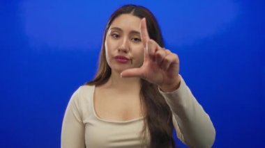Bir kadın, tarafsız bir ifadeyle mavi bir stüdyoda elini yüzünü çerçeveleterek ezik bir işaret hareketi yapıyor; alay ediyor..