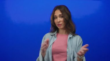 İzole edilmiş mavi bir arkaplana karşı derin düşüncelere sahip genç İspanyol kadın el çırpıyor..