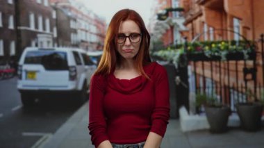 Kırmızı kazaklı kadın arka planda arabalar ve binalarla şehir caddesinde duruyor, gülümsüyor ve kameraya bakıyor, güneşli bir gün, günlük kıyafetler.