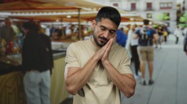 Sakallı genç adam güneşli bir günde alışveriş yapan insanlarla dolu hareketli bir açık hava pazarında rahat duruyor, canlı bir şehir atmosferini yakalıyor..