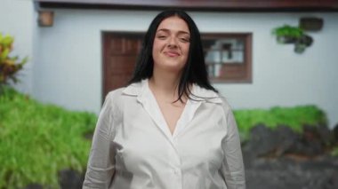 Arka planda bereketli yeşillikle evin dışında el çırpan bir kadın, günlük beyaz gömlekle neşeli bir ifade sergiliyor, huzurlu bir banliyö ortamında güven ve neşe somutlaştırıyor..