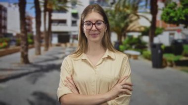 Gözlüklü ve bej gömlekli genç sarışın kadın parlak bir sokakta güven içinde kollarını kavuşturuyor..