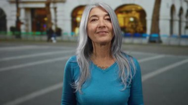 Gri saçlı kadın gülümsüyor ve bir şehir caddesinde işaret ediyor, mavi giyiniyor, arka planda bulanık bir bina var..