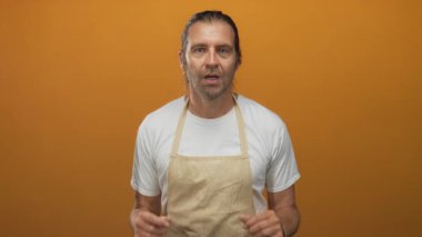 Beyaz tişörtlü, uzun sarı saçlı adam turuncu stüdyo ortamında önlük tutuyor..