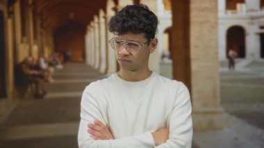 Eski kasaba caddesinde kolları çapraz olan genç İspanyol adam dar kemerli sütun geçidi altında; kafa karışıklığı şüphesi belirsizlik.