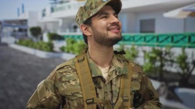 Kamuflaj üniformalı genç İspanyol adam, gülen yüzünü, güneşli bir caddeye bakarken gösteriyor..