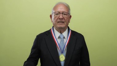 Madalya jestleri ile kıdemli bir adam, izole edilmiş sarı arka plana başparmak kaldırıyor, karışık onay ve hoşnutsuzluk duyguları sergiliyor..